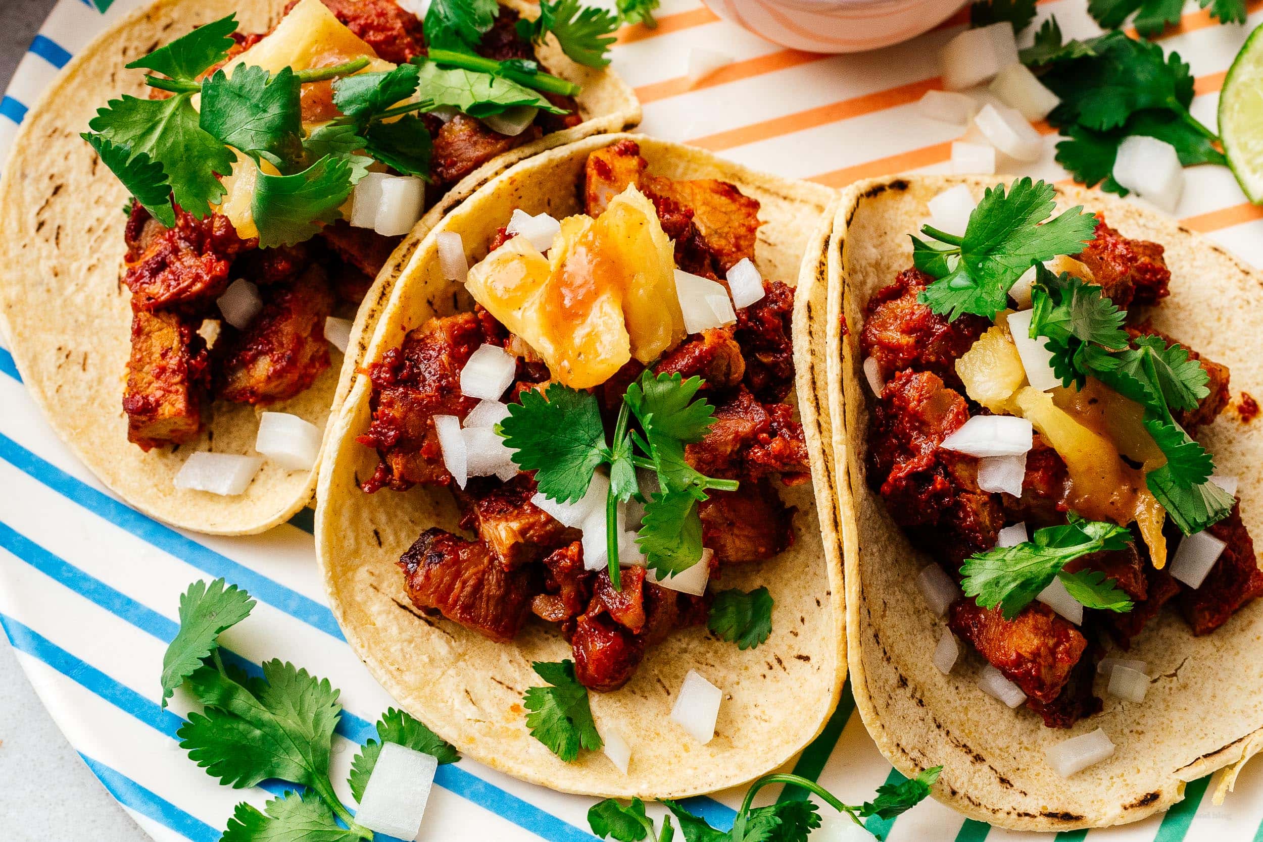 Three al pastor tacos on corn tortillas topped with pineapple, diced onion, and cilantro