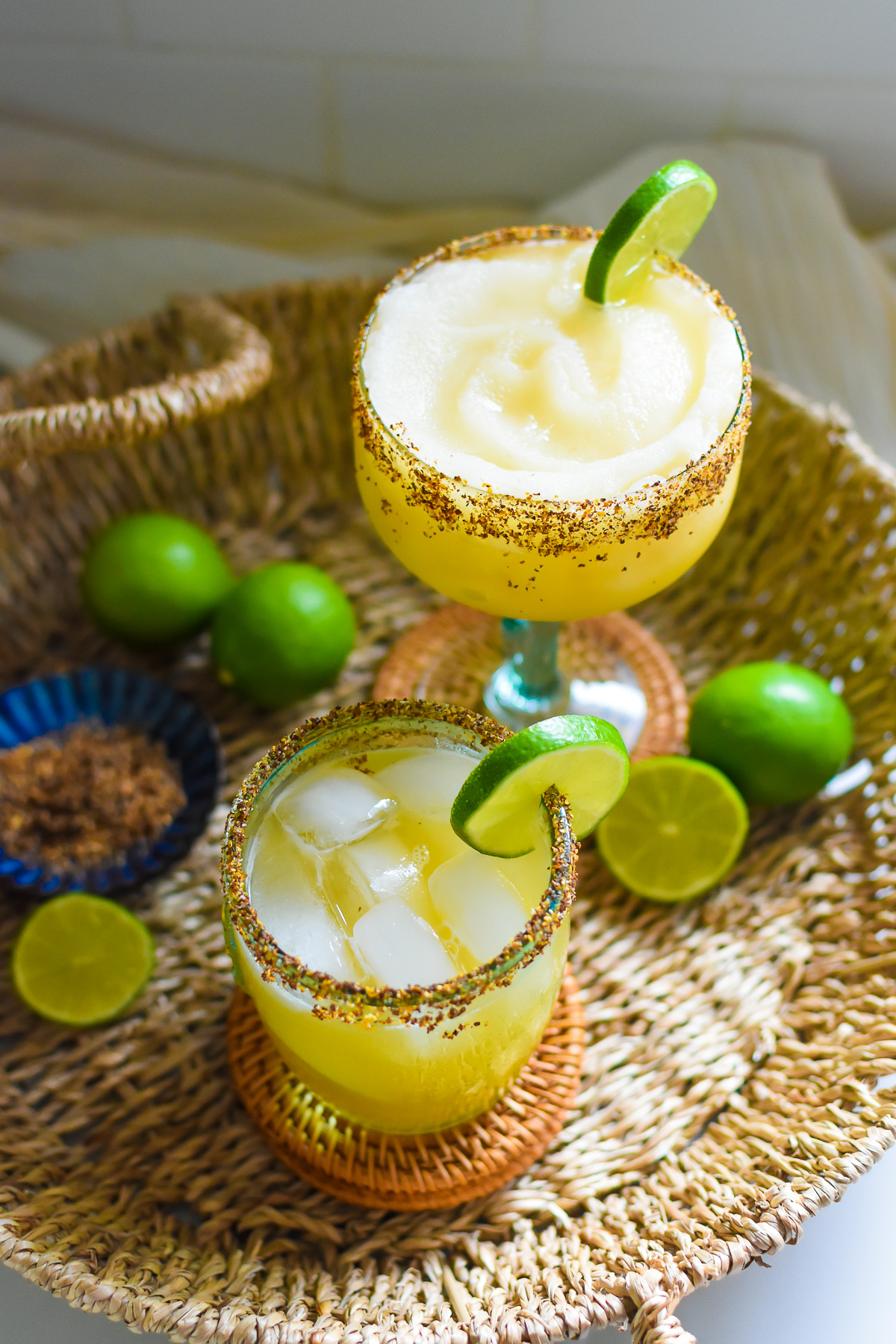 Two margaritas with chili salt rims and lime wedges on a woven tray