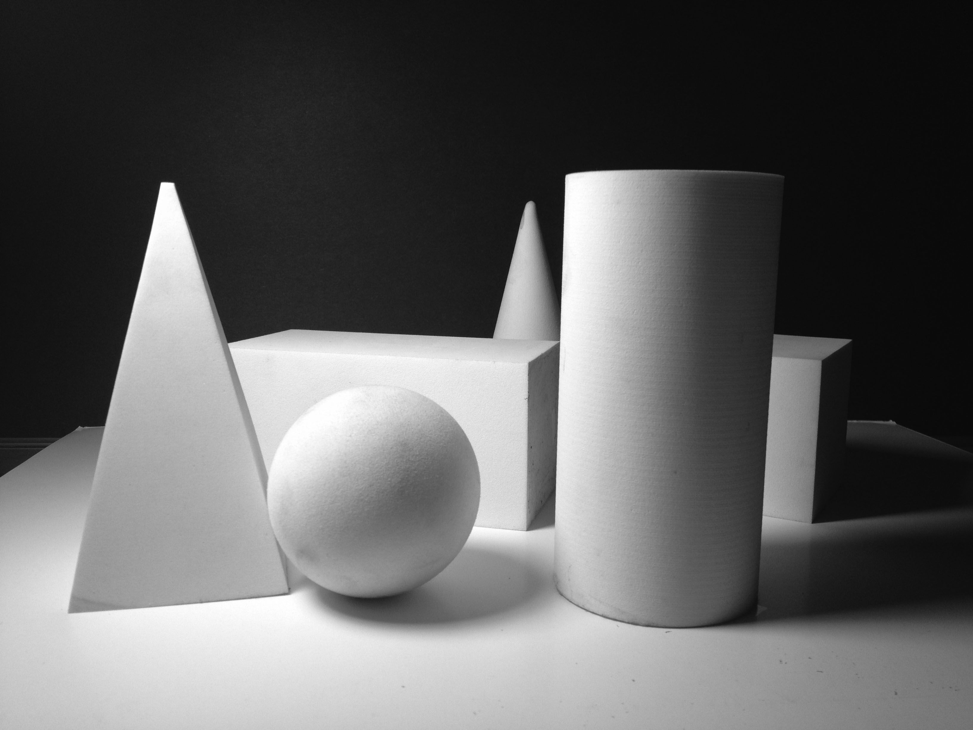 Black and white still life of white plaster solids - cone, sphere, cylinder, cube and pyramid under dramatic light